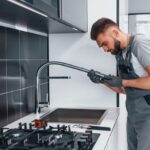Young professional plumber in grey uniform fixing water tap on the kitchen.